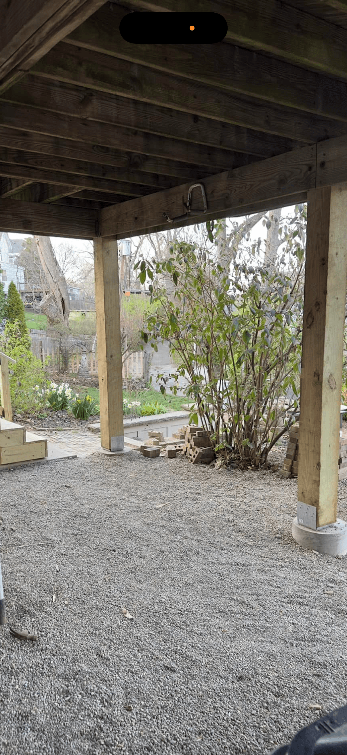Gravel patio under a wooden deck, with garden, daffodils, and bricks beyond.
