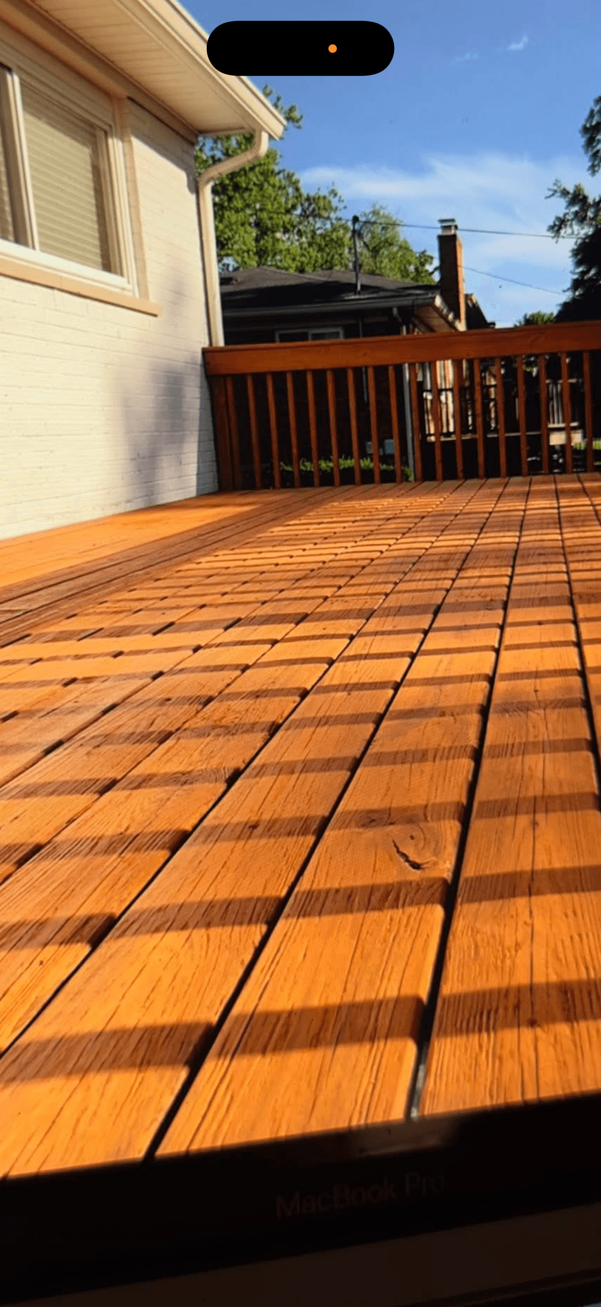 Warm-toned wooden deck with strong striped shadows, white house, and railing under blue sky.